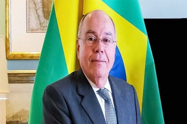 Mauro Vieira in a dark suit and grey tie standing before a Brazilian flag, a framed historical map on the wall behind