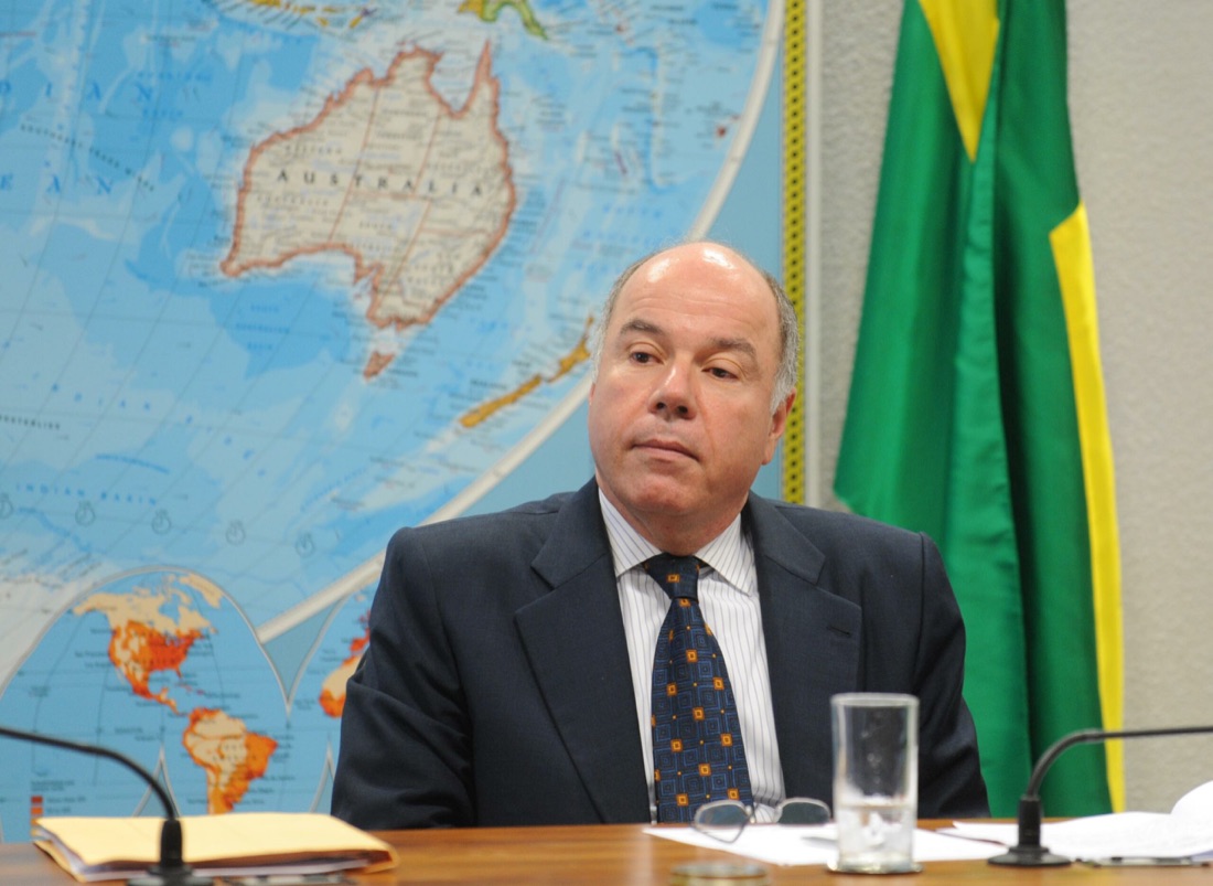 Mauro Vieira seated at a committee table with microphones and a glass of water, a large world map and a Brazilian flag behind him