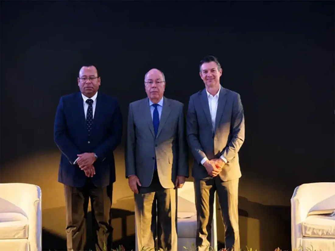 Mauro Vieira standing on a stage between two other men in suits, white armchairs to either side