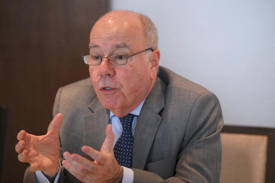Mauro Vieira, wearing glasses and a grey suit, gestures with both hands while speaking at a table