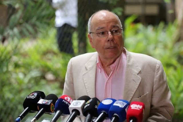 Mauro Vieira in a light jacket and pink shirt speaking to reporters at a cluster of broadcast microphones outdoors, greenery behind