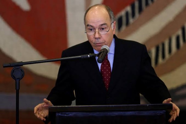 Mauro Vieira speaking at a lectern microphone with arms extended, a large red and white tapestry behind him